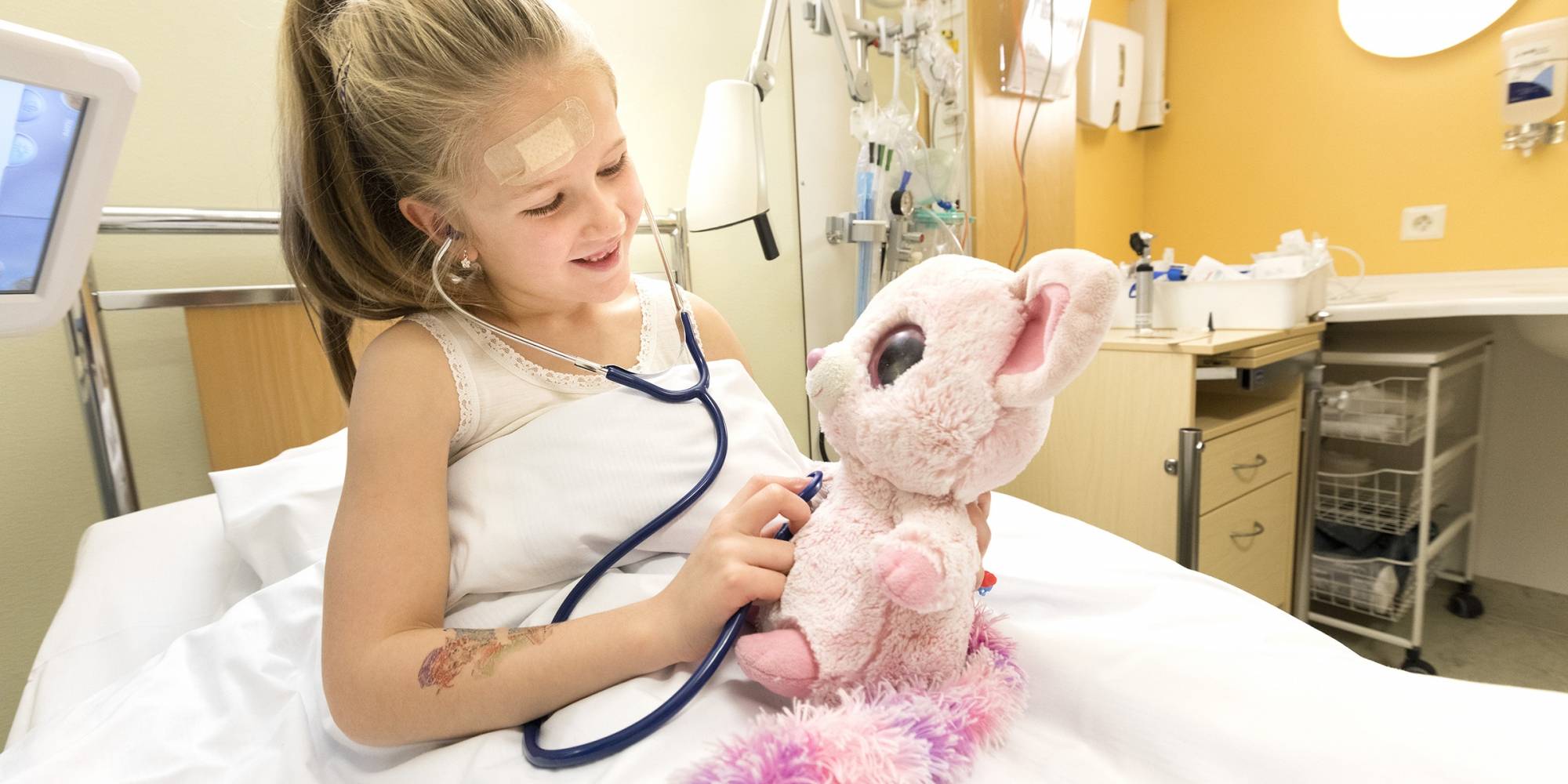 Jente i sykehusseng lytter på hjertet til kosedyret sitt med et stetoskop. Foto: Gorm Kallestad / NTB og Sonja Balci